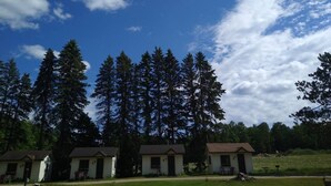 A row of small, single-story cabins with a grassy lawn in front, surrounded by tall pine trees under a partly cloudy sky. - Cabin #9 -" Flag-up " (Cadillac)