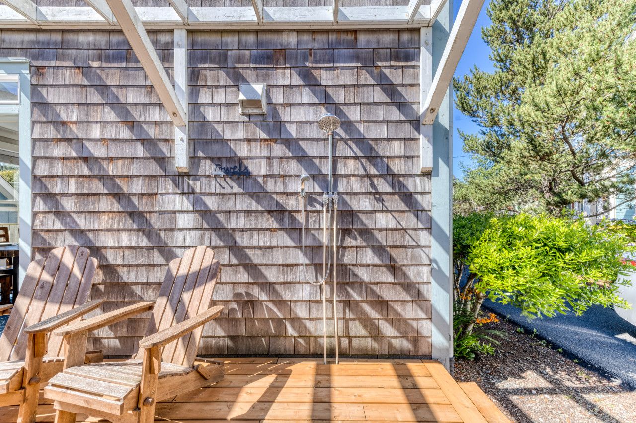 A wooden deck with Adirondack chairs, a metal showerhead, and a wooden wall with shingles.