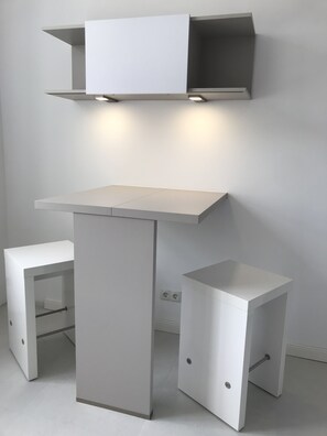 Ein schlicht eingerichteter Raum mit weißem Schreibtisch und passendem Hocker, einem Regal darüber sowie einer Wandsteckdose.
