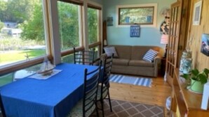 A room with a dining table, chairs, a sofa, and a window with a view of greenery. - Maggies on the Bayou, close to Hiking, Beaches, Boating, Golf (Ludington)
