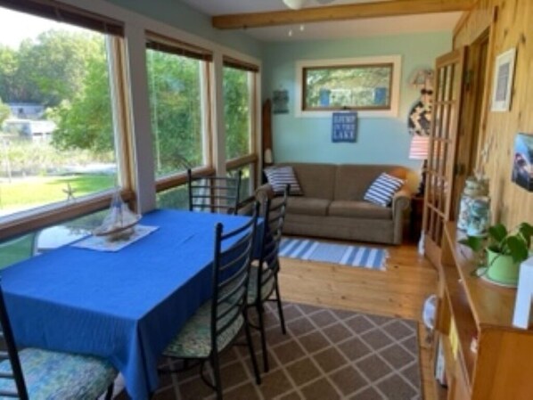 A room with a dining table, chairs, a sofa, and a window with a view of greenery.