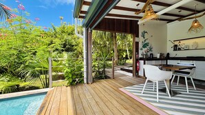 Una terraza de madera con piscina, un comedor con sillas blancas y una cocina con lámpara colgante.