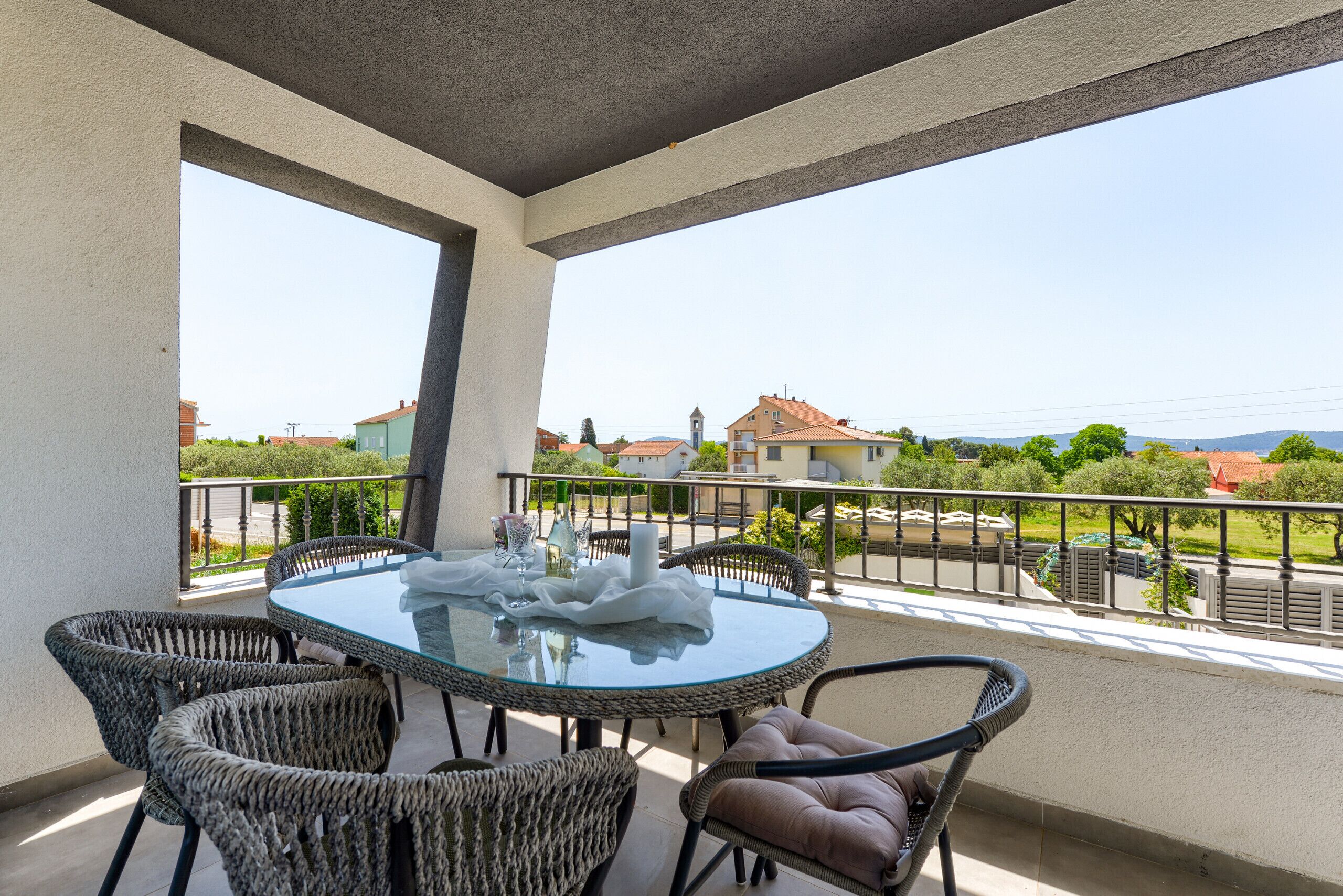 A balcony with a glass table and wicker chairs, overlooking a residential area with houses and a church.