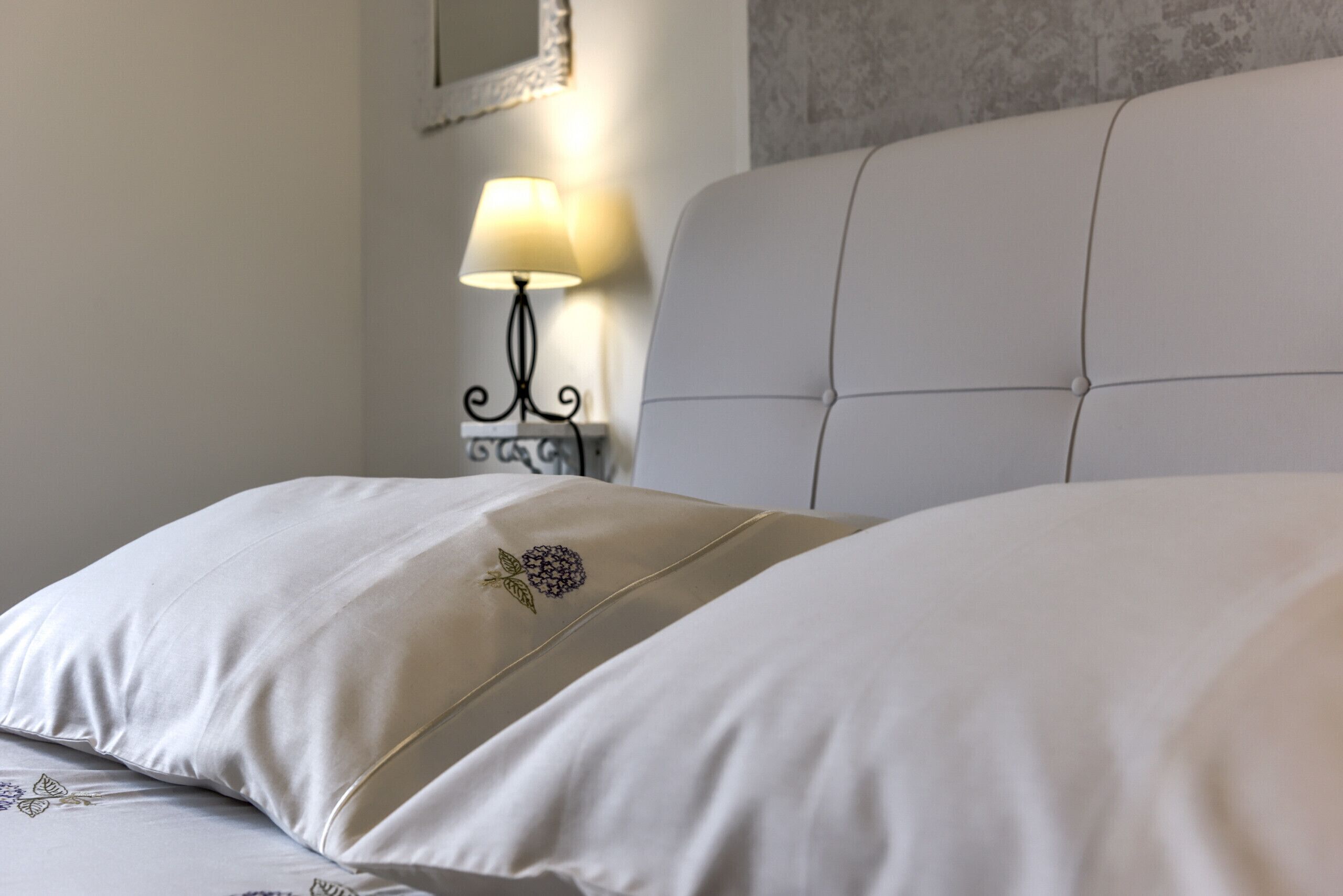 A neatly made bed with a floral embroidered pillow and a lamp on a bedside table.
