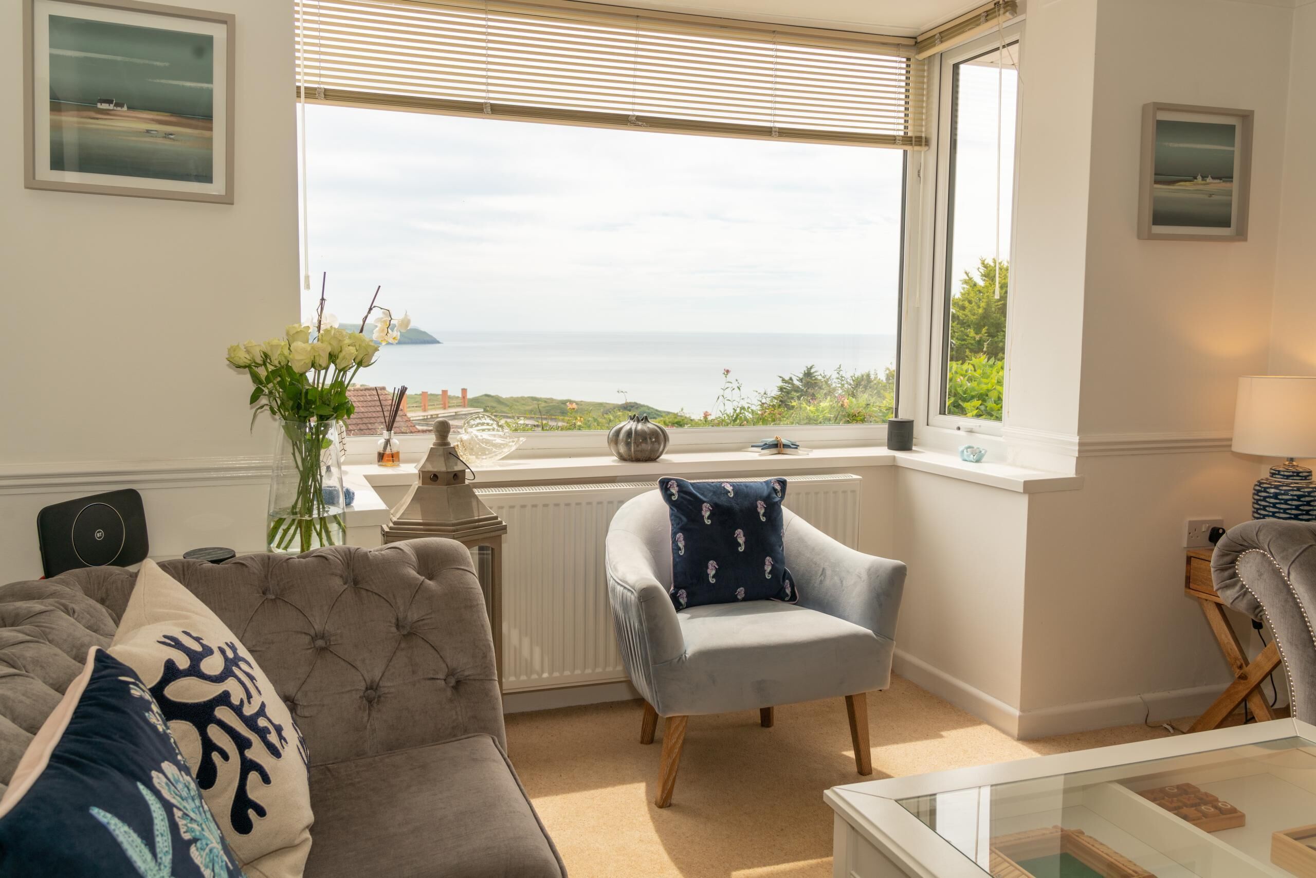 A living room with a view of the sea, a grey sofa, a blue armchair, and a glass coffee table.