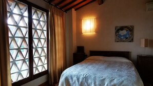 Une chambre avec un plancher en bois, un lit recouvert d’une courtepointe blanche, une table de chevet, une lampe fixée au mur et une fenêtre ornée d’un motif en treillis.