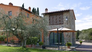 Un bâtiment en pierre avec une entrée dotée d’une porte en verre, des places assises à l’extérieur et une pergola surmontée d’un parasol.