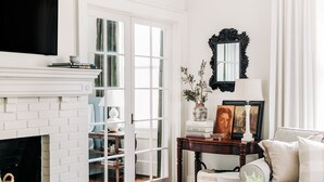 A living room with a fireplace, a television, a sofa, and a dining table with a tray of snacks. - Designer Retreat in Quaint Historic District w Front Porch Dining under the oaks (Greenville)