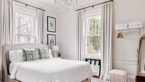 A bedroom with a large bed, a chandelier, a window with curtains, a bedside table, and a mirror. - Designer Retreat in Quaint Historic District w Front Porch Dining under the oaks (Greenville)
