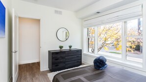 Een slaapkamer met een groot raam, een donkere dressoir, een ronde spiegel en een bed met een grijs en zwart gestreept dekbed.
