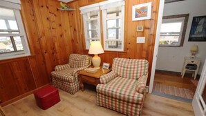 A cozy room with wooden walls, a plaid sofa set, a small table with a lamp, and a red ottoman. - "The Hammock Cottage" (Kill Devil Hills)