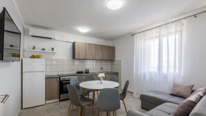 A modern kitchen with a dining table and chairs, a grey sofa, and a window with sheer curtains. - Click Here - Get Promo Price By Inquiry - Apartments Nenna With 3 Bedrooms (Mravince)