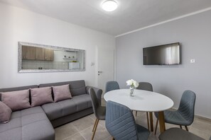 A modern living room with a grey sectional sofa, a round dining table with chairs, a flat-screen TV mounted on the wall, and a large mirror above the sofa.