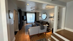 A modern living room with a sofa, a coffee table, a TV, and a large mirror. There is a staircase leading to another level. - Downtown Darling & Convention Center Home (Salt Lake City)