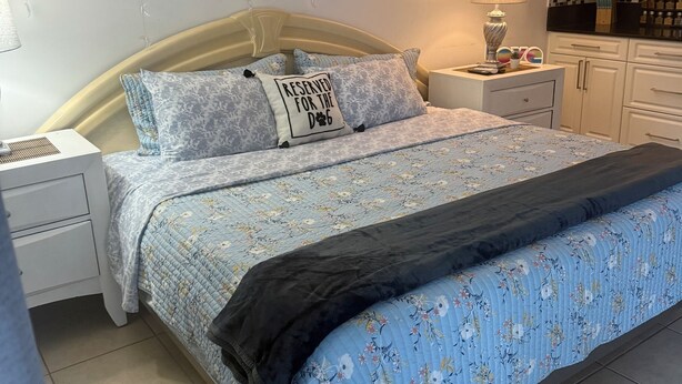A bedroom with a bed, nightstands, a lamp, a wall clock, and a kitchen area with cabinets.