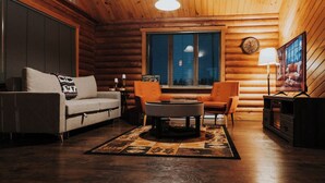A log cabin interior with a sofa, two armchairs, a coffee table, a fireplace, and a large window with blinds.