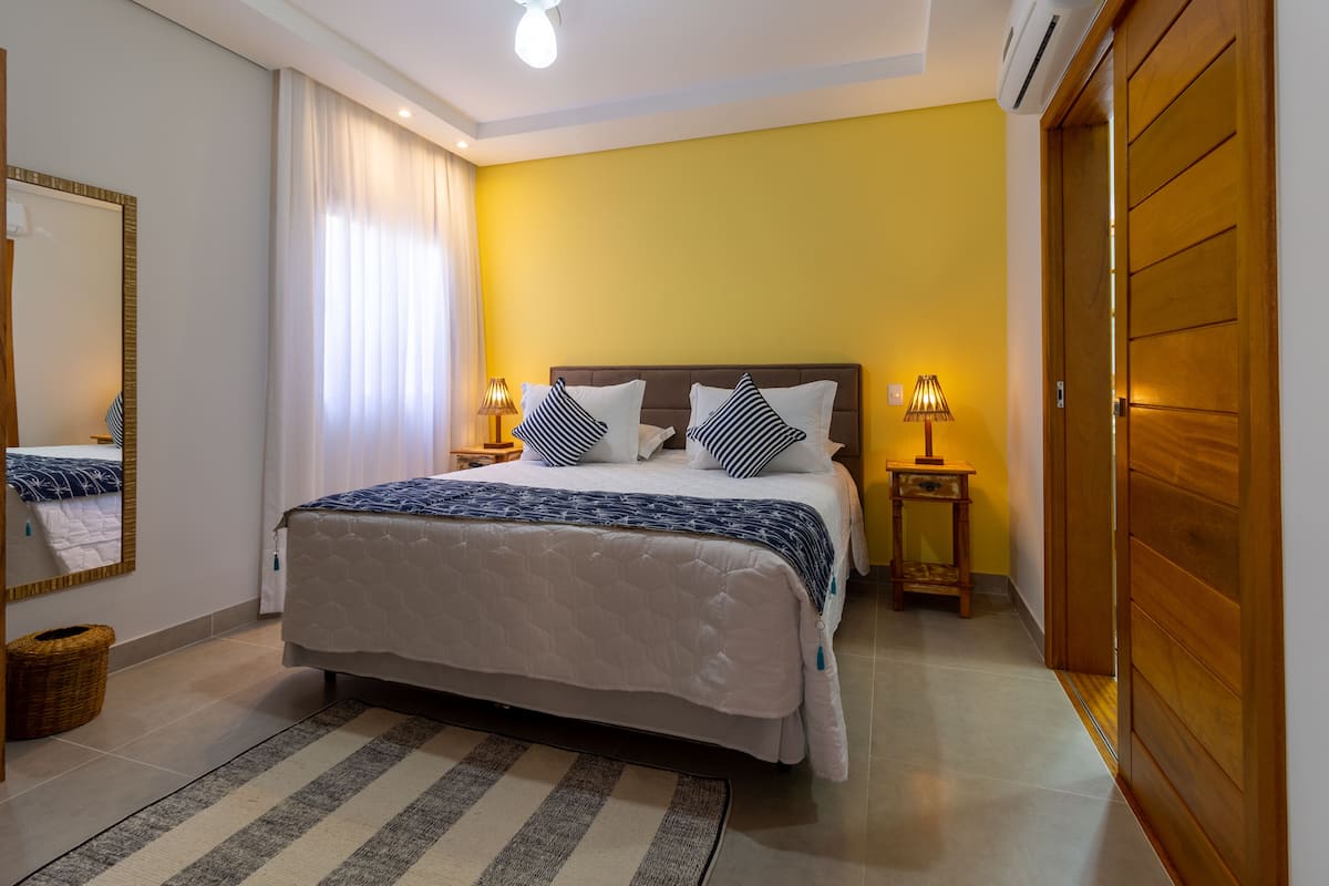 A hotel room with a large bed, two bedside tables, a mirror, a wooden door, and a yellow wall.