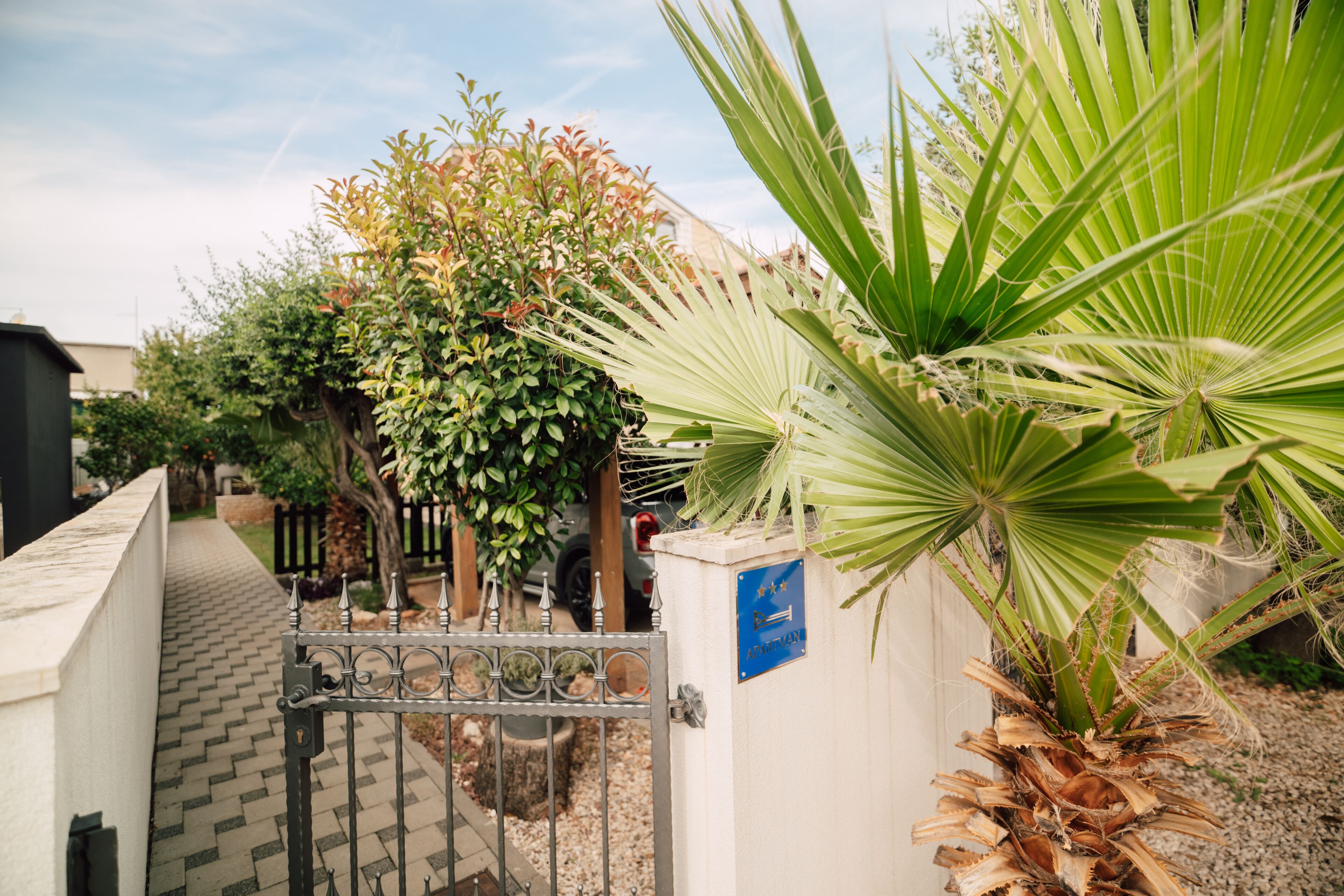 A gated entrance with a pathway leading to a building, surrounded by various plants and trees.
