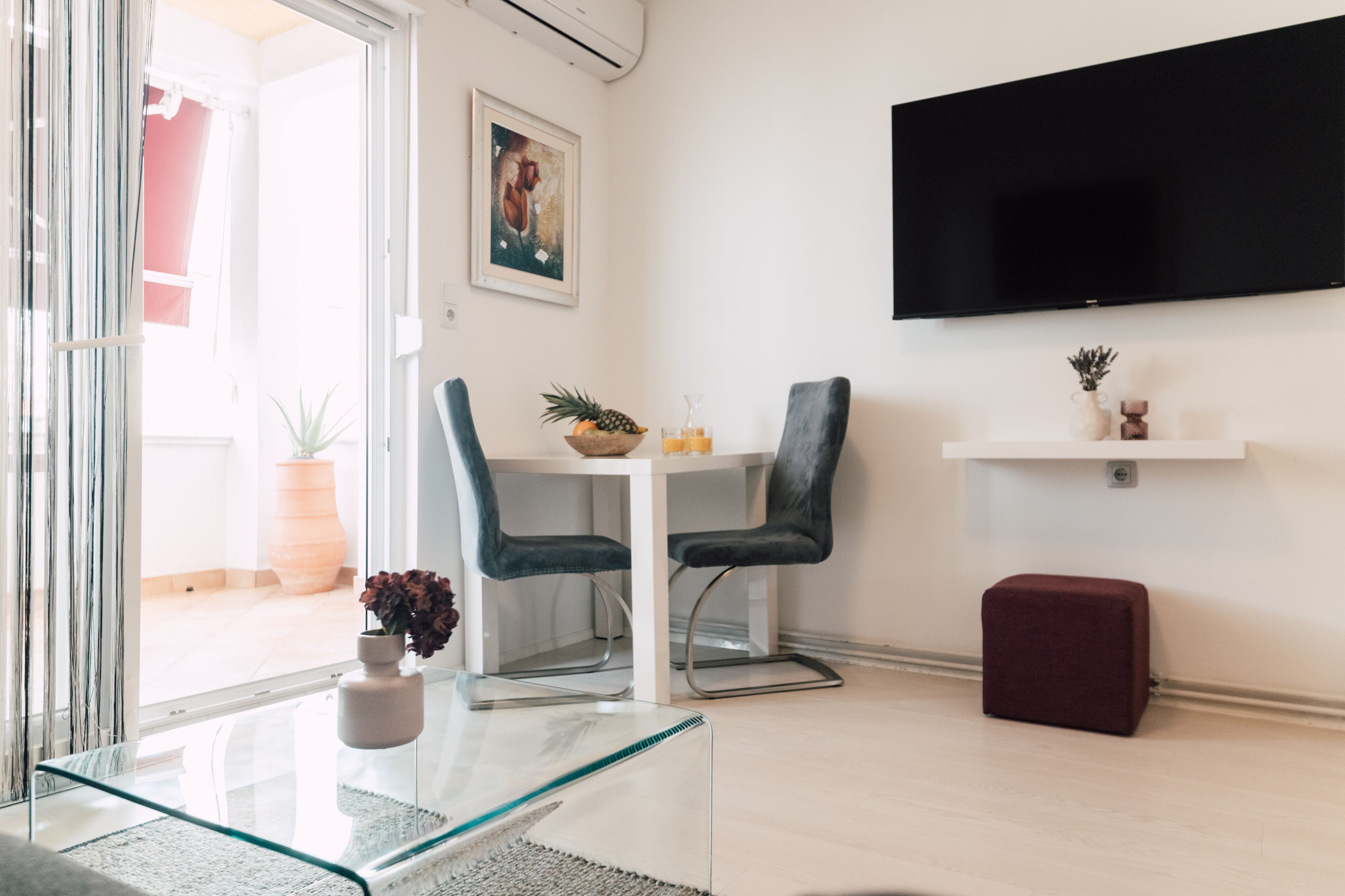 Een moderne woonkamer met een glazen salontafel, twee donkere fauteuils, een witte tafel, een wandgemonteerde televisie en een klein rode poef.