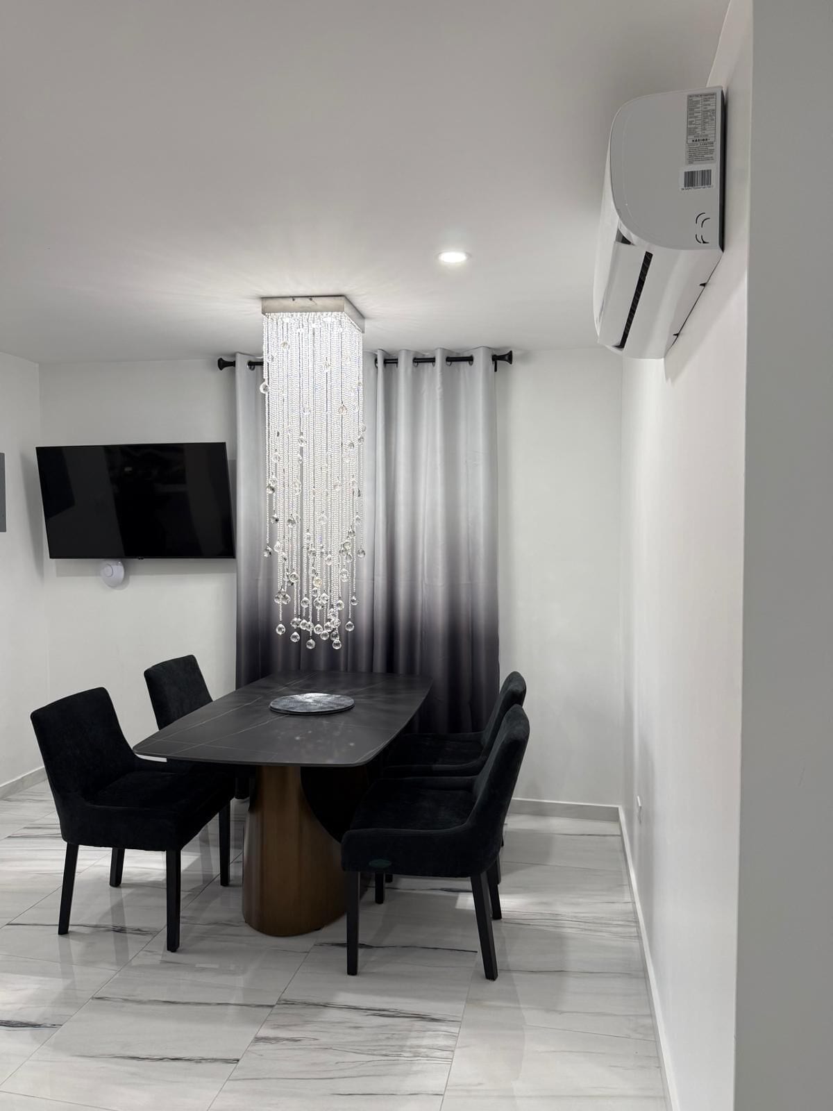 A modern dining room with a rectangular table, black chairs, a chandelier, and a mounted TV.