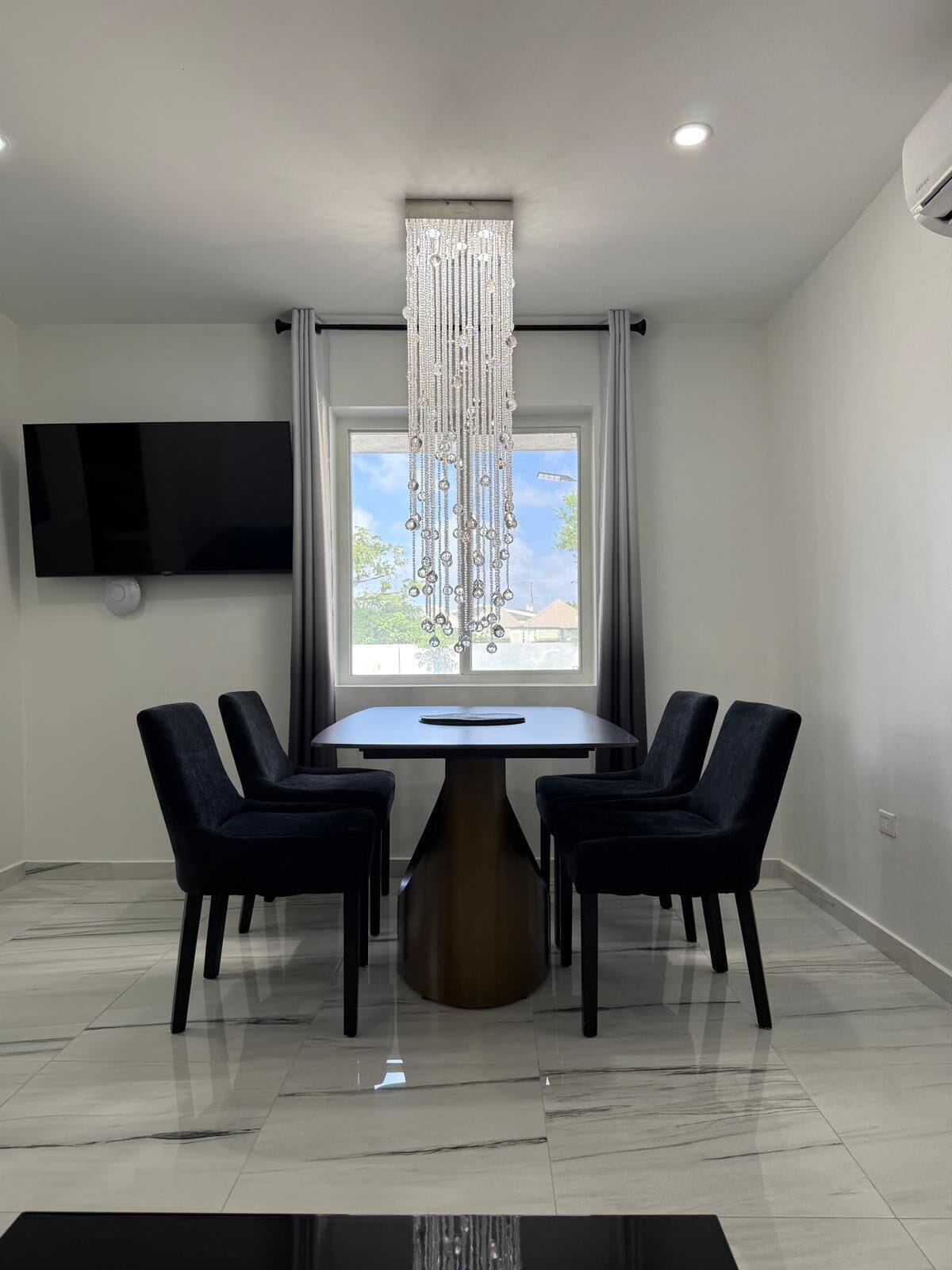 A modern dining room with a rectangular table, four chairs, a chandelier, a flat-screen TV, and a window with curtains.