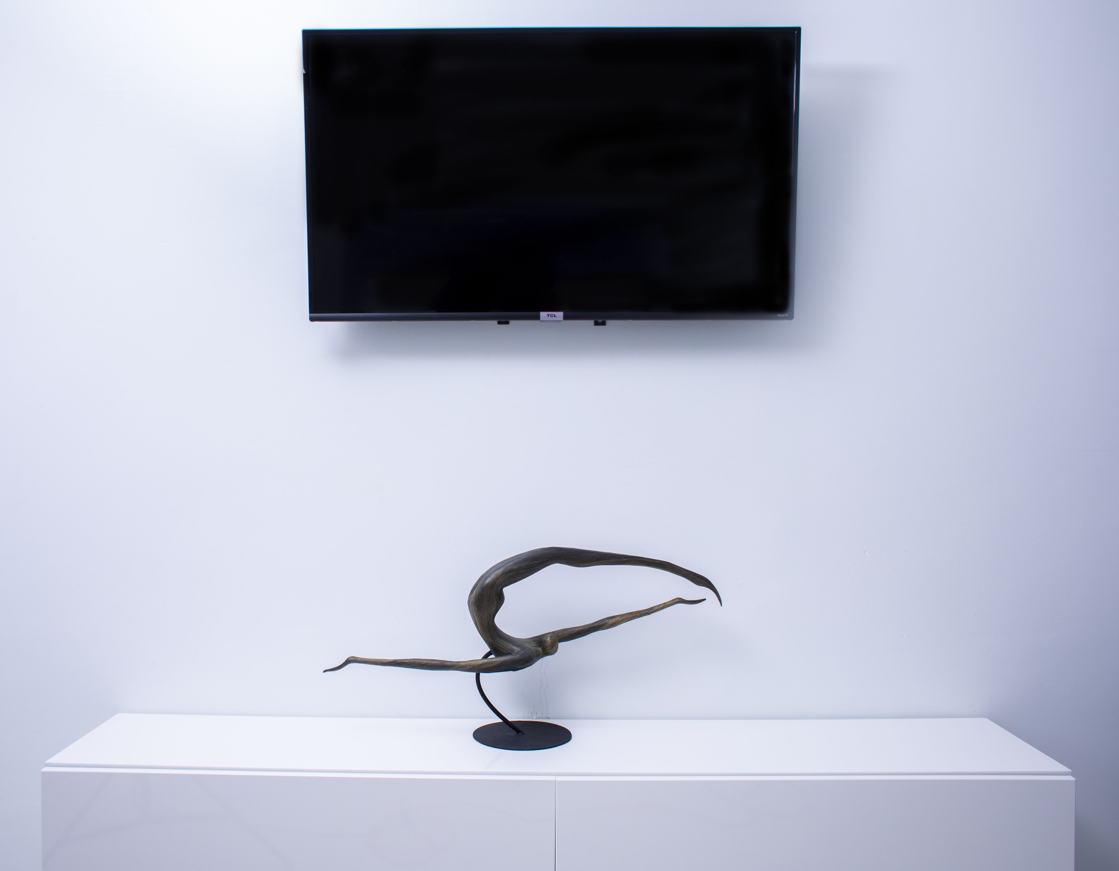 Un televisor de pantalla plana sujeto a una pared blanca sobre un mueble blanco con una escultura de metal encima.