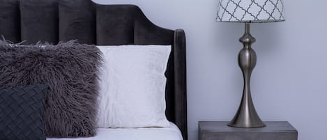 Una cama con colcha blanca y almohada gris mullida, una lámpara de metal con pantalla estampada y una mesita de noche de madera con cajón.