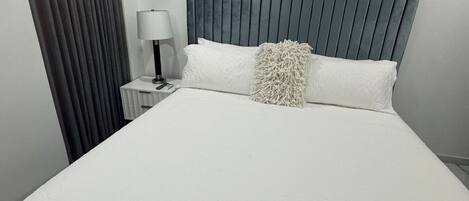 Un dormitorio moderno con una cama grande, una mesita de noche, una lámpara y un ventanal con cortinas.