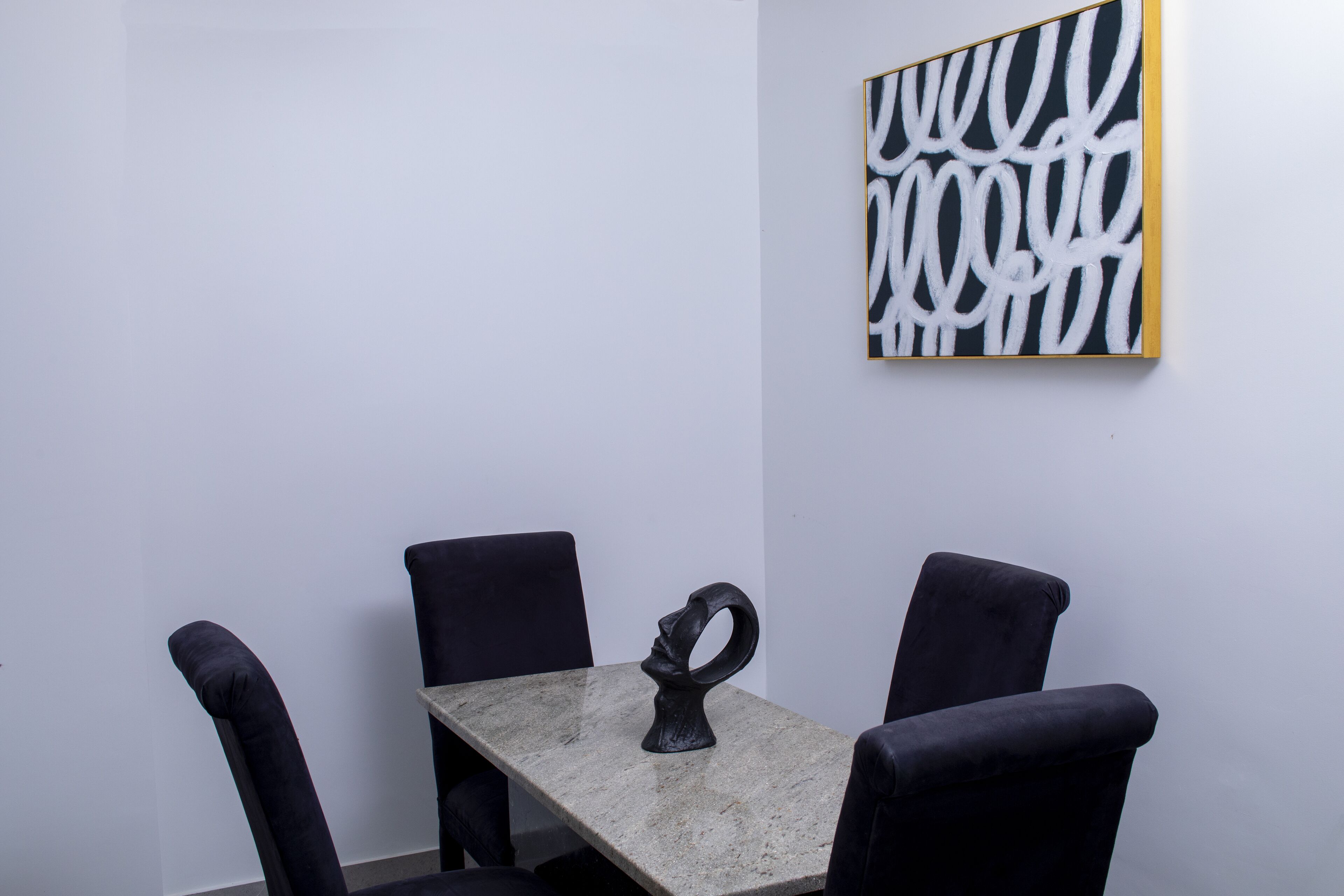 A minimalist room with a table and four chairs, a black abstract painting on the wall, and a sculptural object on the table.
