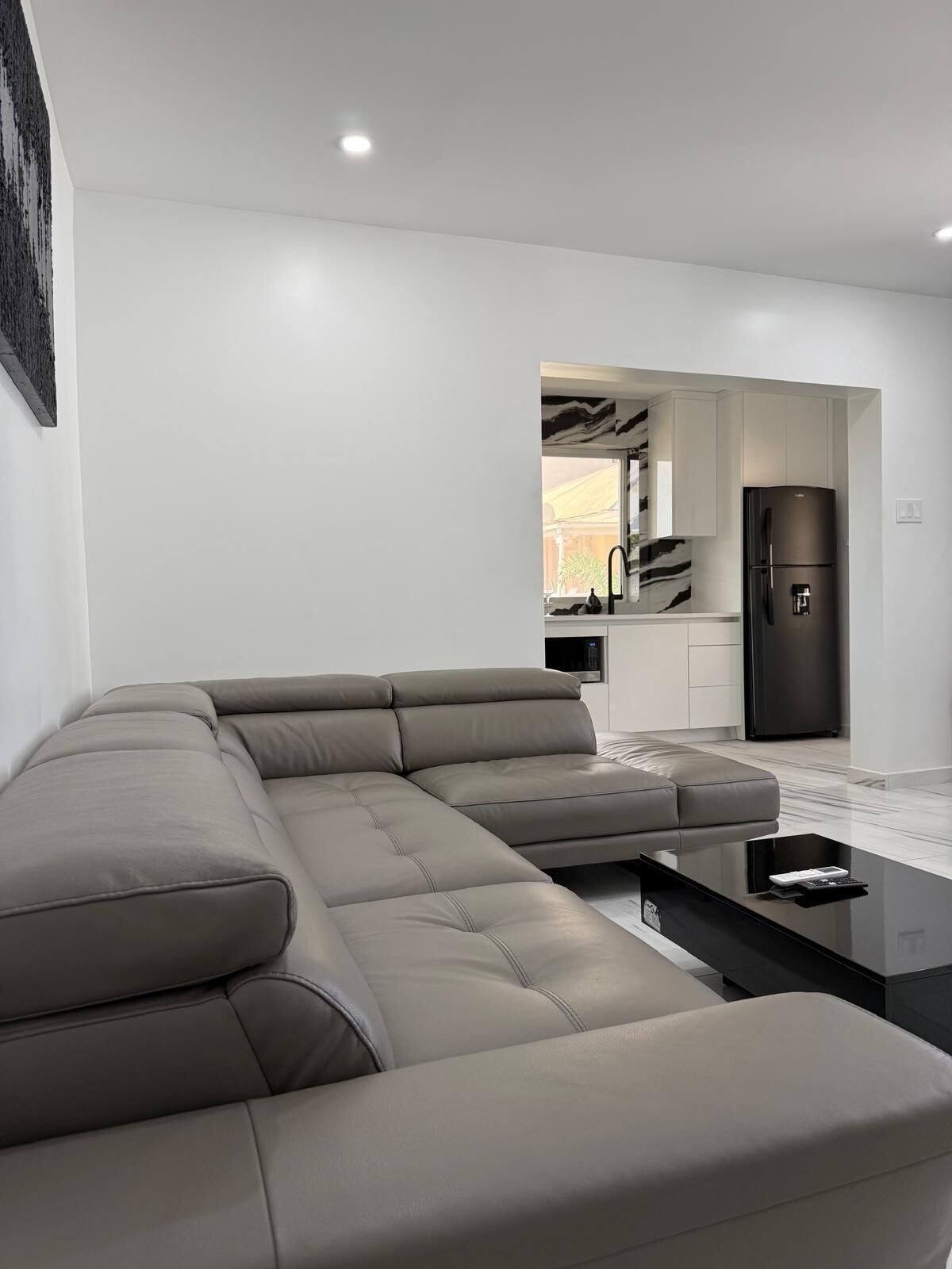 A modern living room with a grey sectional sofa, a black coffee table, and a kitchen area with a black refrigerator.