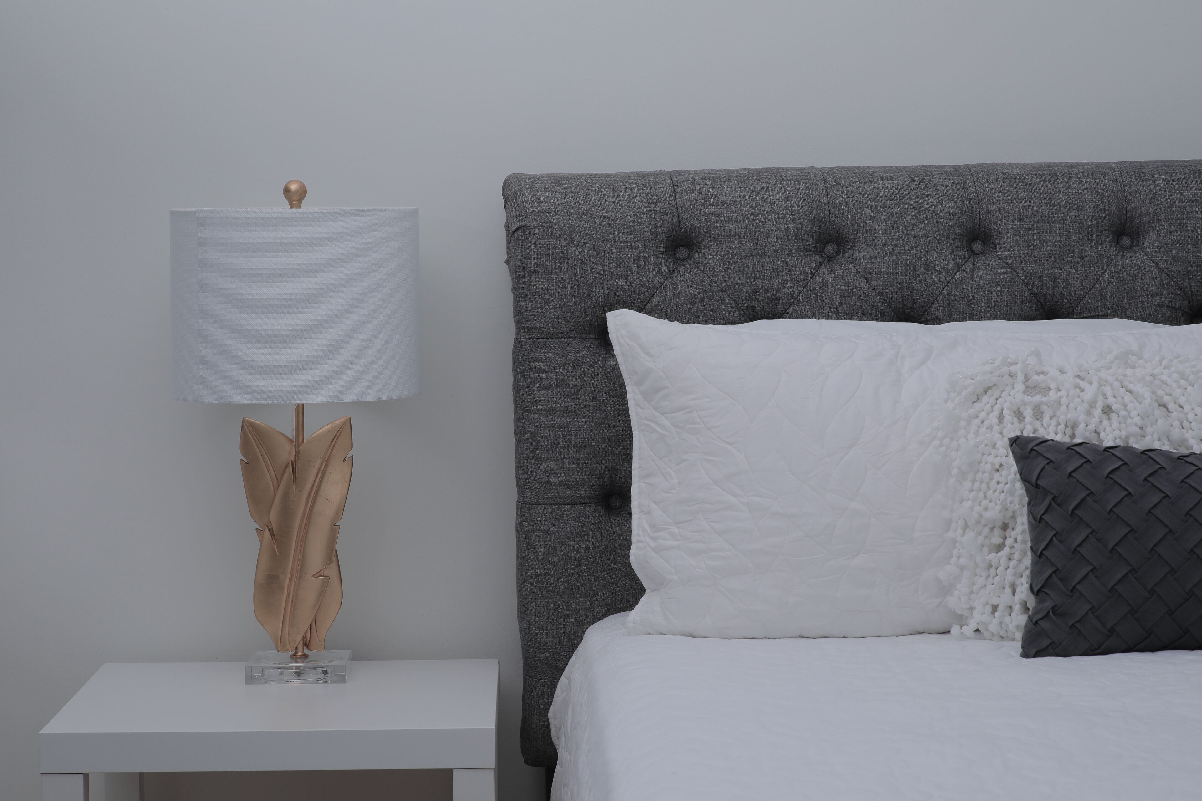 Un lit bien fait, avec une tête de lit rembourrée de couleur grise, des oreillers blancs et noirs, et une table de chevet blanche agrémentée d’une lampe et d’un objet décoratif.