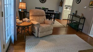 A living room with a large armchair, a dining table with chairs, a kitchen area, and a ceiling fan.