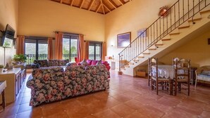 A spacious living area with a large floral sofa, a dining table with chairs, a staircase, and a view of the outdoors through large windows. - Montion Jimera de Libar by Ruralidays (Málaga)