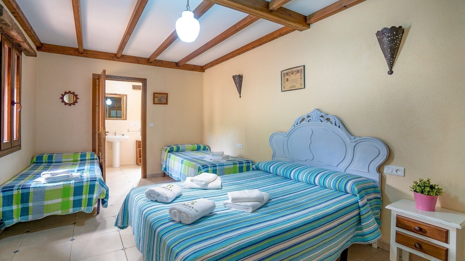 A bedroom with two single beds and a larger bed, all with striped bedding. There is a small bedside table with a plant and a wall-mounted light fixture.