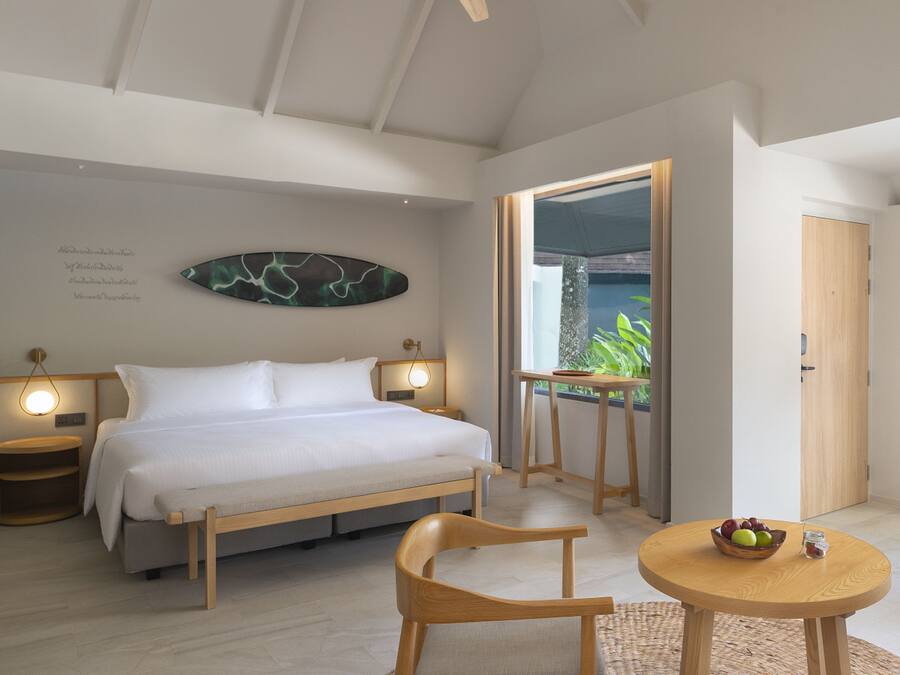 Una habitación de hotel moderna con cama, muebles de madera, una mesita con fruta y vistas al verdor por la ventana.
