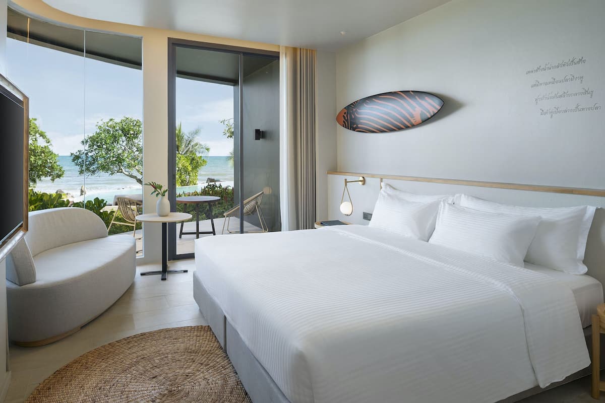 A modern hotel room with a large bed, a seating area with a chair and table, and a view of the ocean through a glass door.