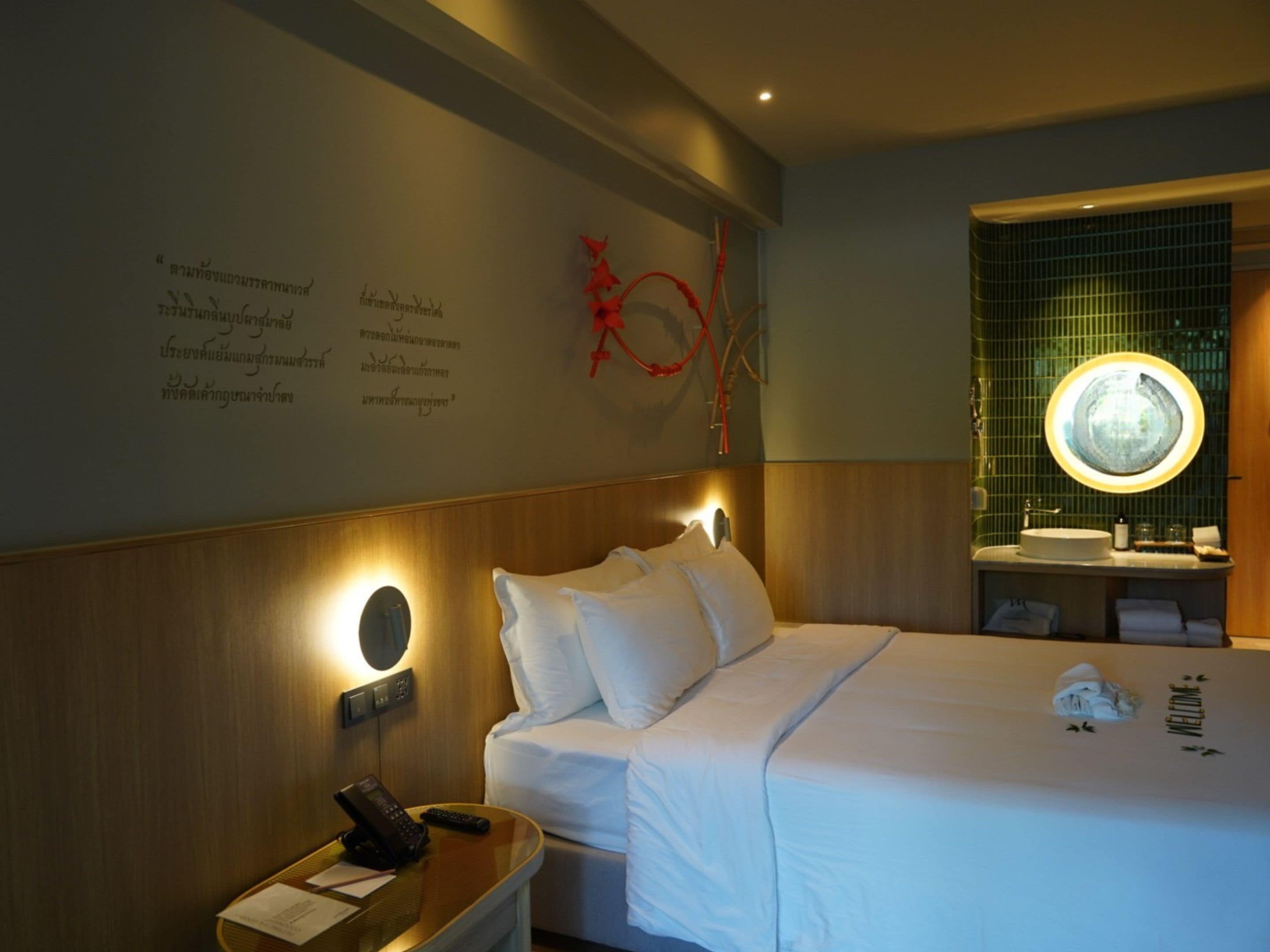 A hotel room with a bed, bedside table, wall-mounted phone, and a round mirror above the sink.