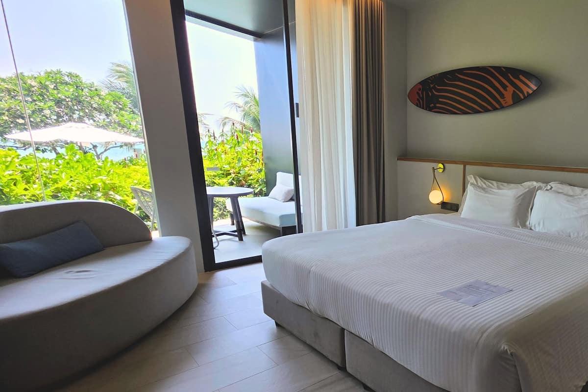 A hotel room with a bed, a sofa, a small table, and a view of greenery outside.