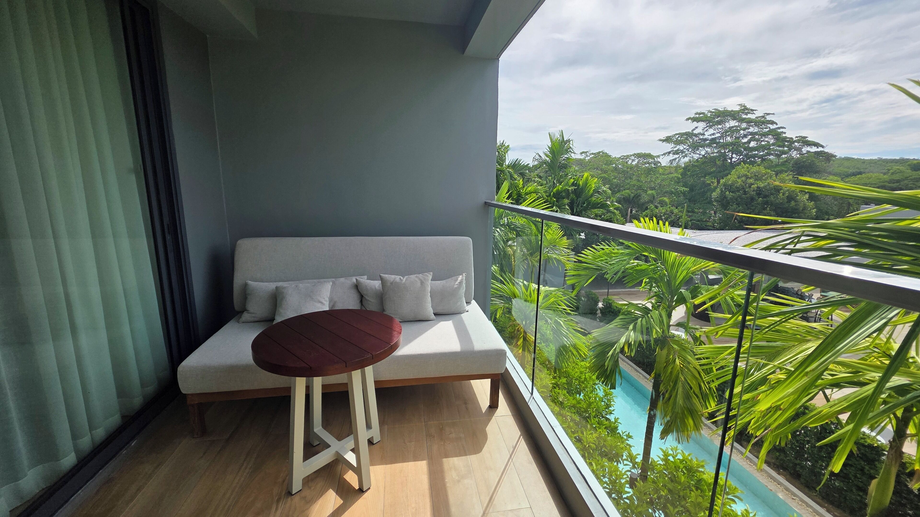 A balcony with a sofa and a small table, overlooking a swimming pool and lush greenery.