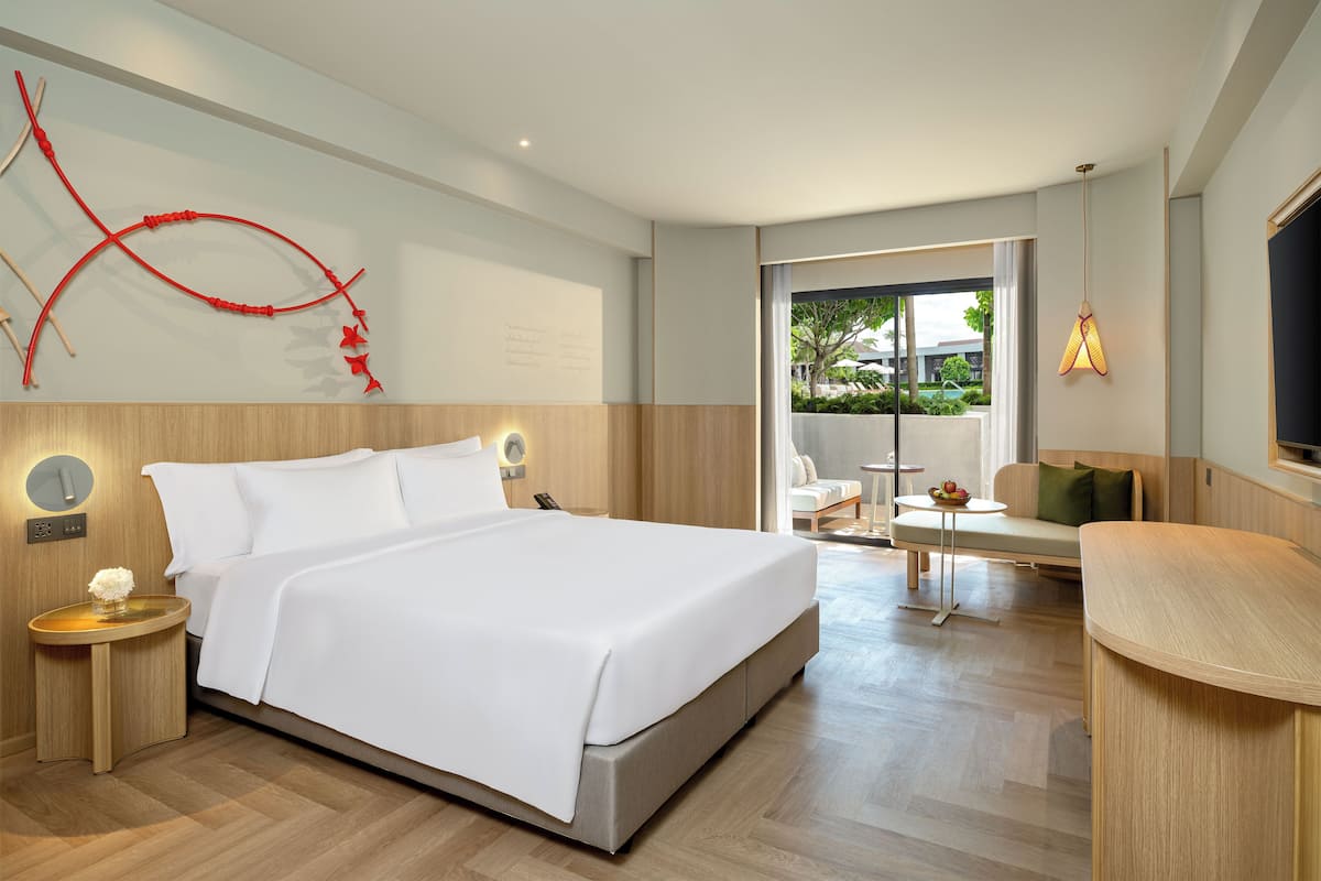 A modern hotel room with a large bed, a wooden headboard, a wall-mounted artwork, a round bedside table with a lamp, and a view of the outdoors through a glass door.