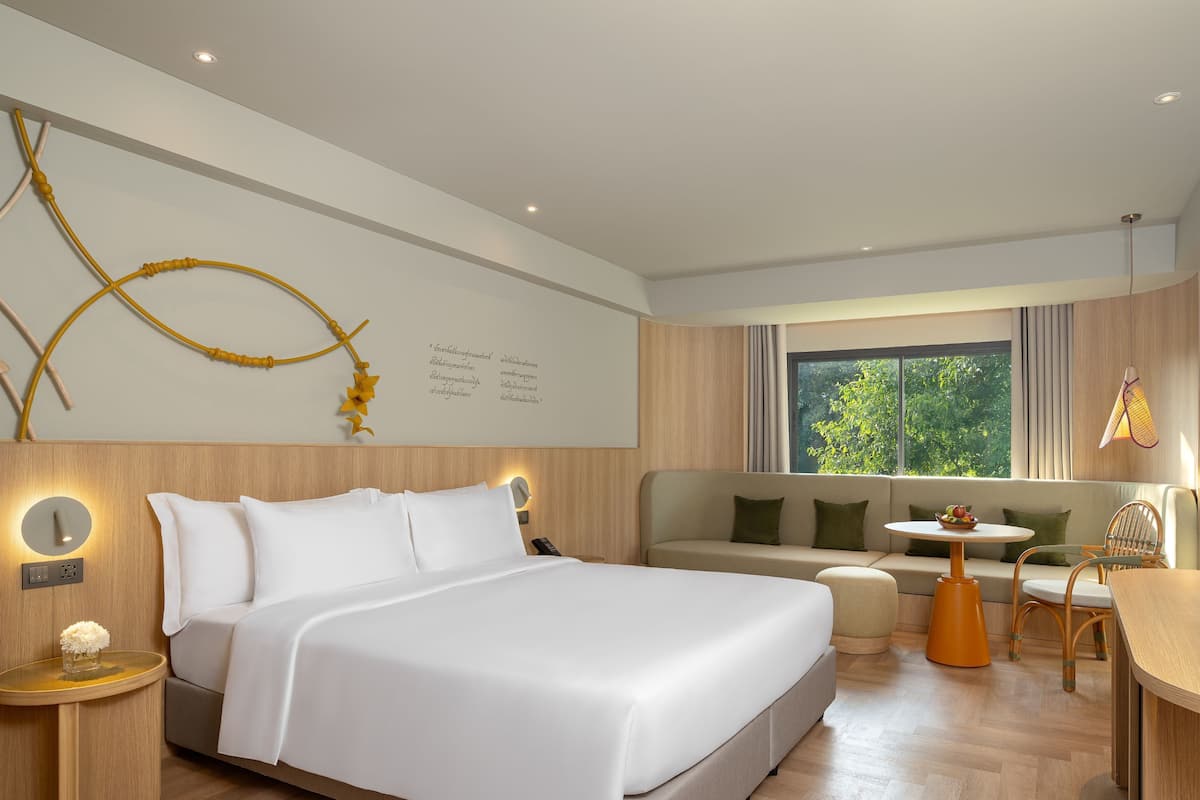 A modern hotel room with a large bed, a sofa, a small table, and a view of greenery outside.