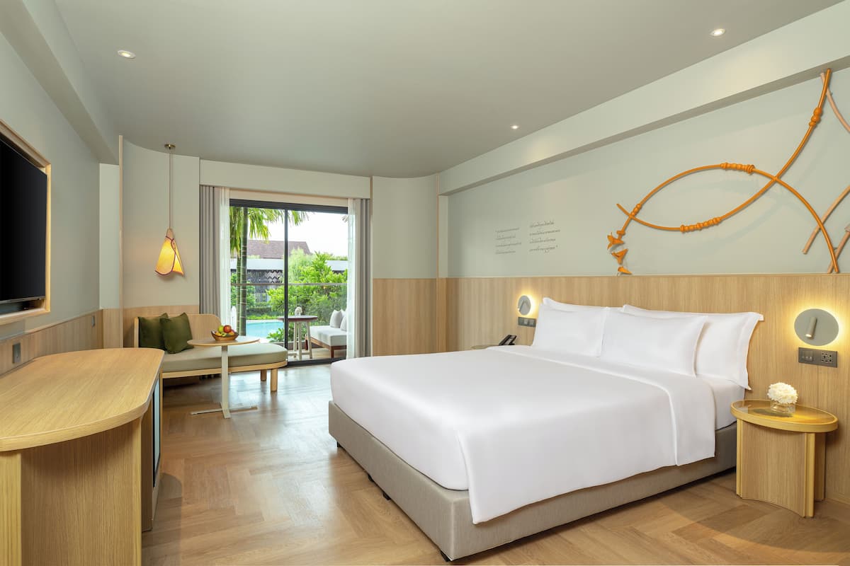 A modern hotel room with a large bed, a desk, and a seating area with a view of greenery outside.