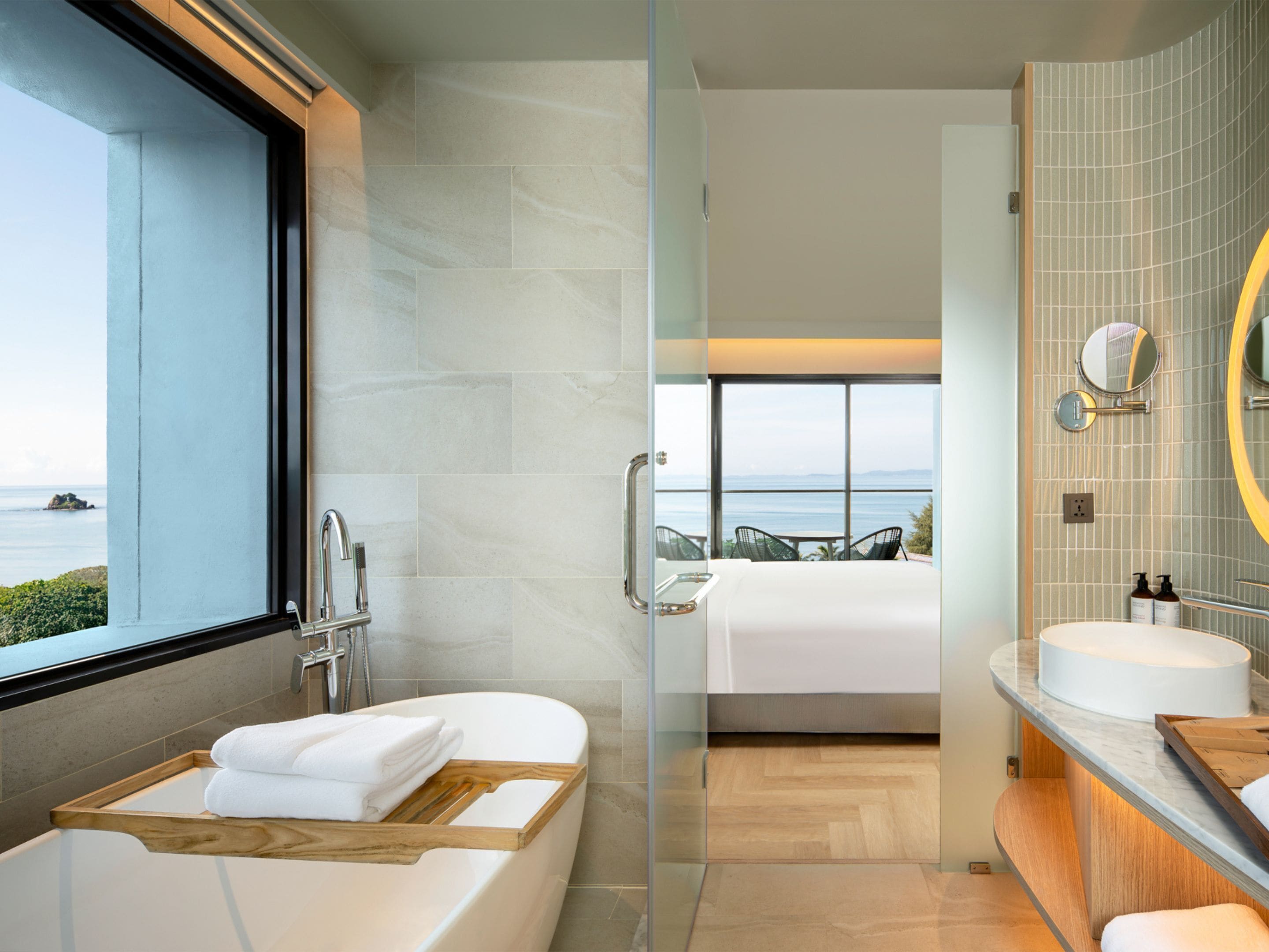 A modern bathroom with a bathtub, a shower, and a large mirror. There is a view of the sea through the window.