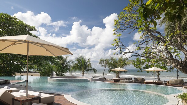 Zona de piscina con sillones y sombrillas, con vista a una playa y al océano.