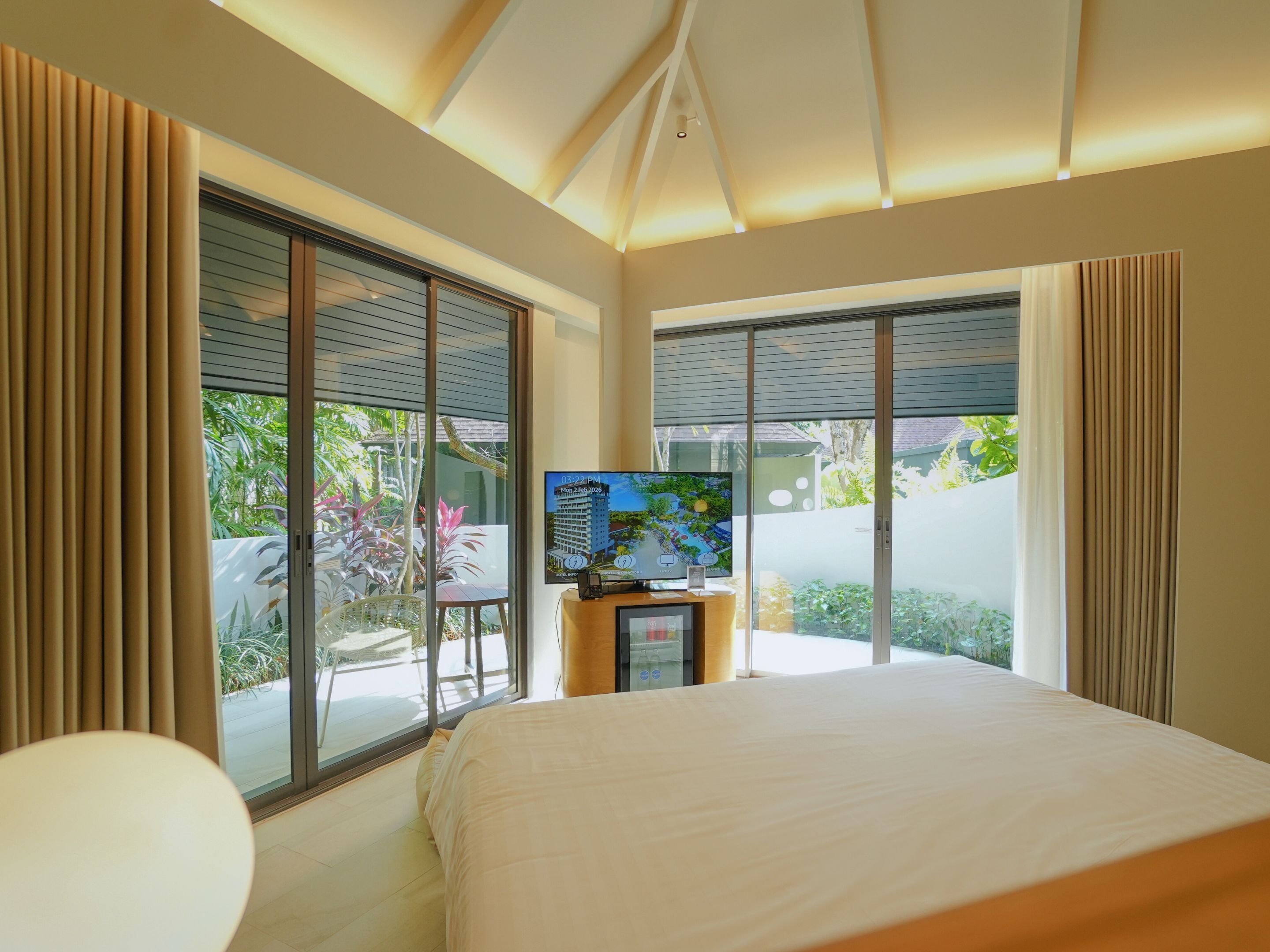 A bedroom with a bed, a TV on a stand, and large windows with curtains.