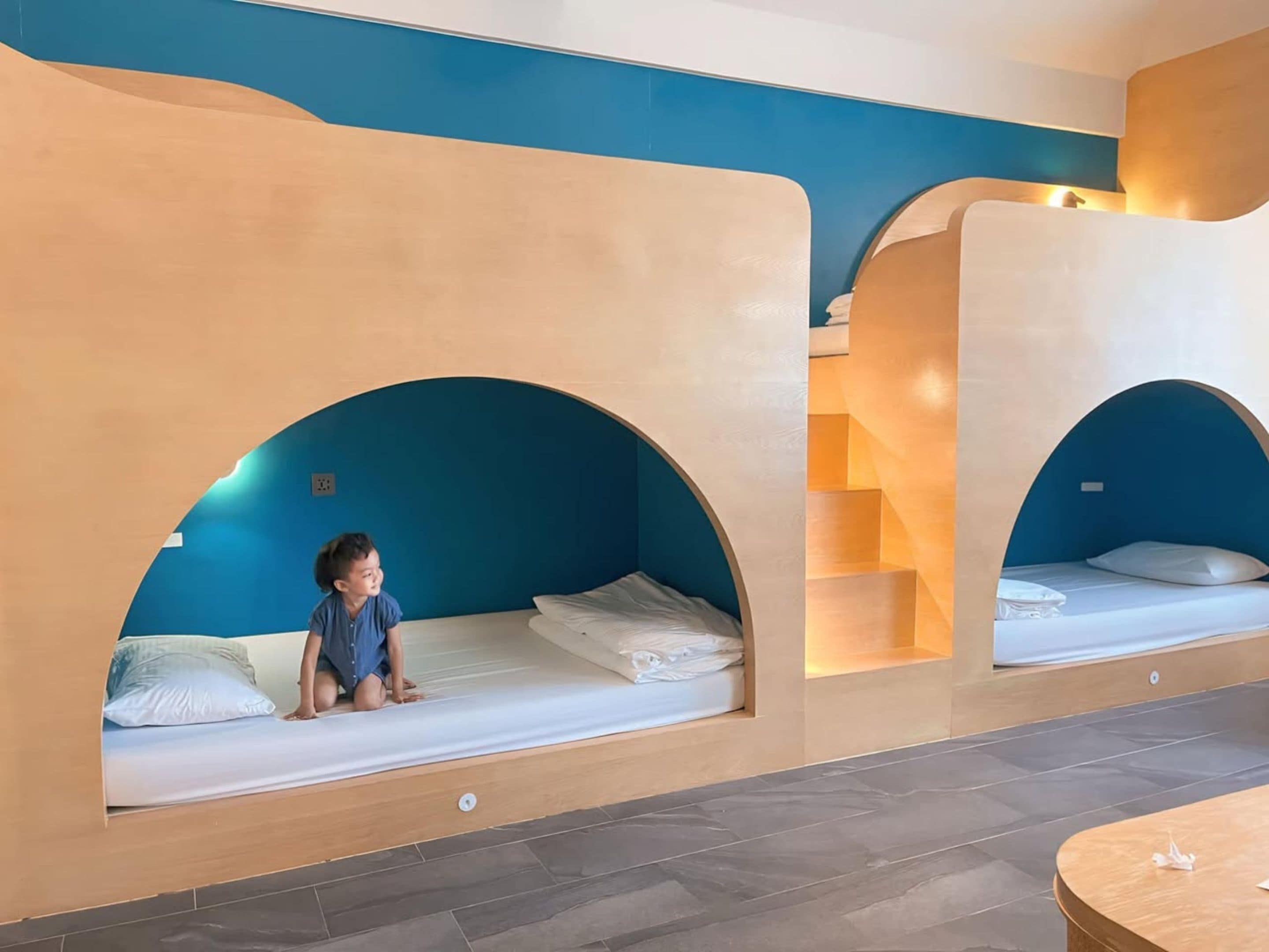 A child sitting on a bed in a room with blue and beige walls, wooden floors, and a staircase.