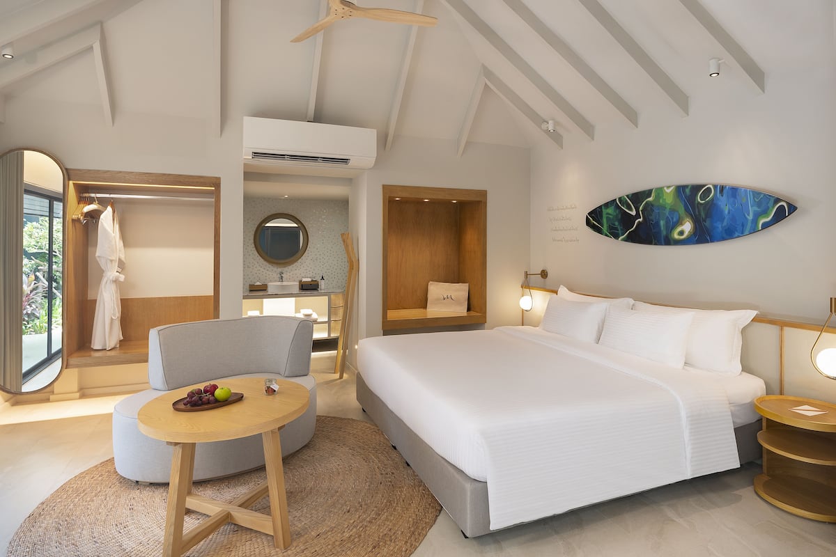A modern hotel room with a large bed, a round dining table, a seating area, and a large mirror.