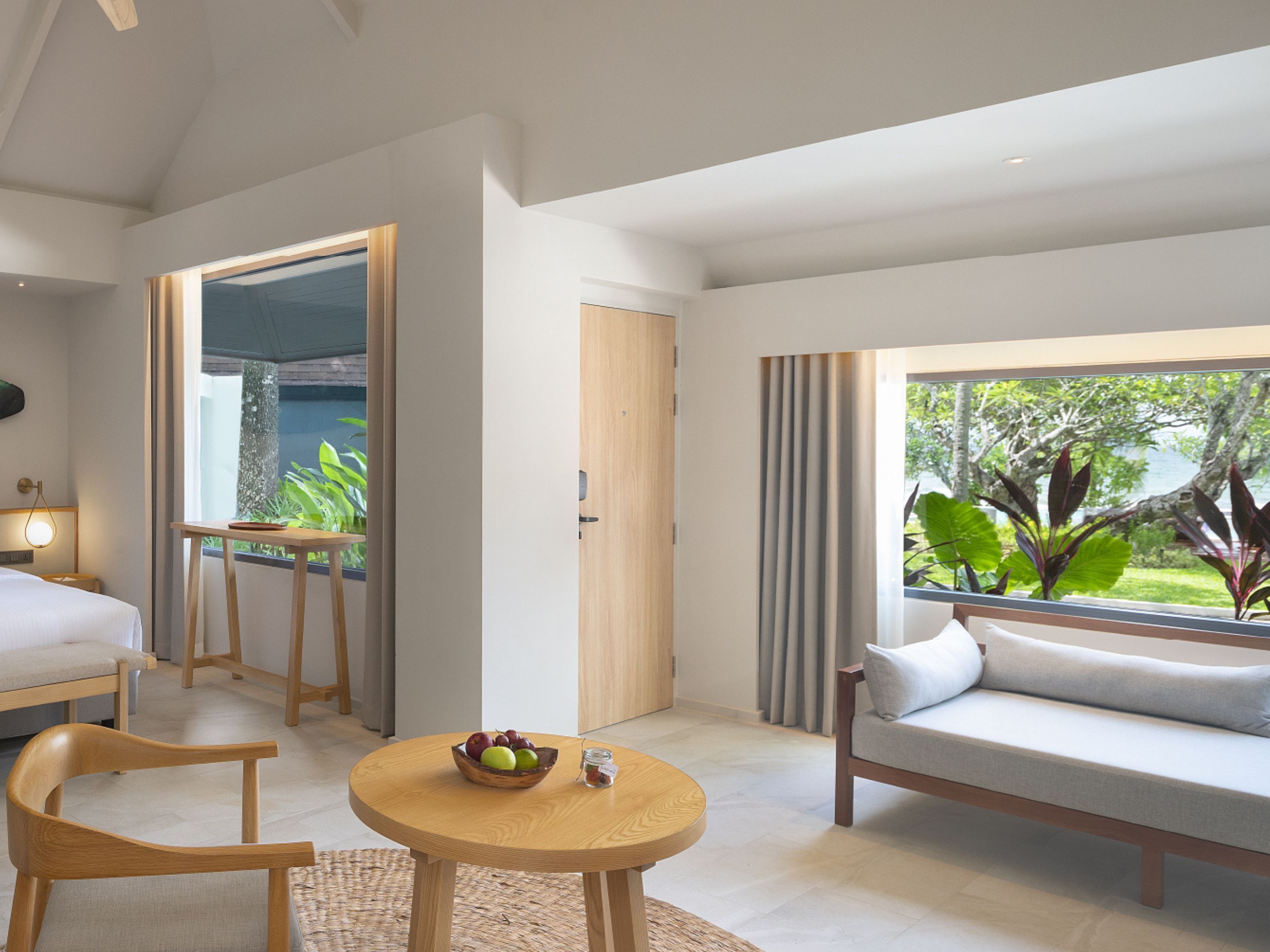 Habitación de hotel moderna con cama, sofá, mesa de comedor y frutero, y vistas a zonas verdes y agua.