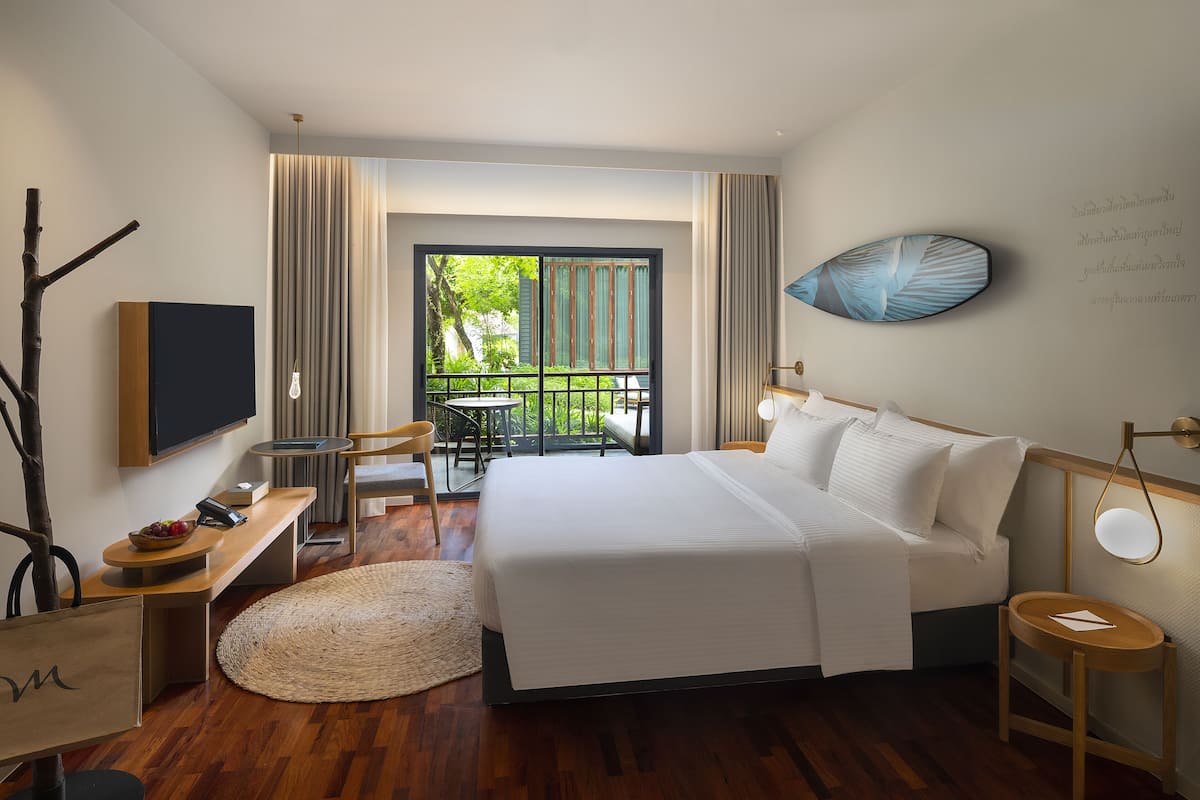 A hotel room with a bed, a desk, a chair, a television, and a balcony with a view of greenery.