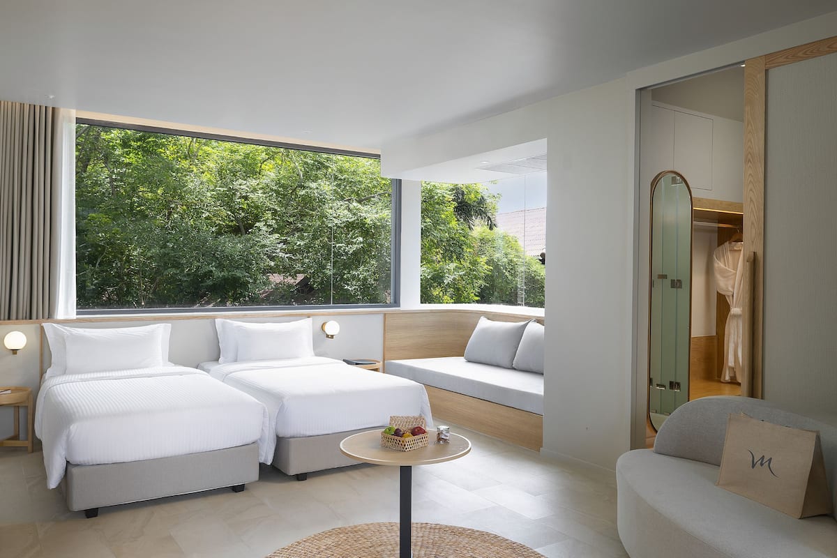 A modern hotel room with two beds, a sofa, a round table, and a view of greenery outside.